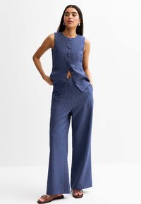 Gilet bleu marine à fines rayures et pantalon large. Le gilet a un col rond et cinq boutons. Tissu lisse avec une coupe ajustée.