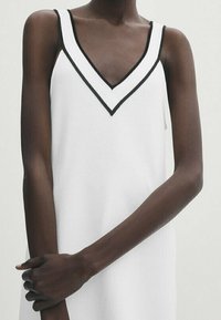 Vestito bianco senza maniche con una scollatura a V profonda, caratterizzato da un bordo nero lungo il collo. Il tessuto appare liscio con una texture a maglia.