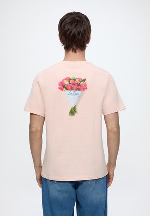 Mand iført lyserød T-shirt med en buket af røde og orange blomster indpakket i papir med teksten "Les Deux" trykt på ryggen.