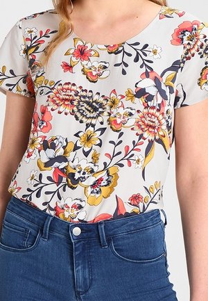 Femme portant un haut blanc à manches courtes avec des motifs floraux colorés, associé à un jean en denim bleu taille haute.