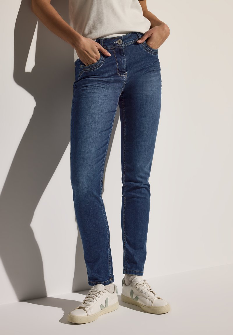 Jeans en denim bleu foncé avec une coupe slim, cinq poches et une fermeture à bouton, associés à des baskets blanches avec des accents vert clair.