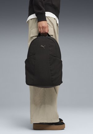Person, der holder en sort tekstureret Puma-rygsæk i toppen, iført beige cargobukser og sorte sko med gummisåler.