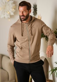 Beige zip-up hoodie lavet af blødt stof, med en forlommer, justerbare snore og et subtilt logo på brystet. Kombineret med sorte bukser.
