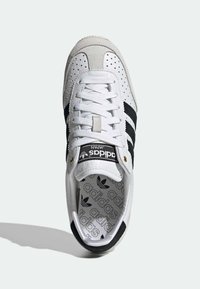 Bílá kožená teniska s černými pruhy a perforovanými detaily. Obsahuje štítek na jazyku a vzorovanou stélku. Zaoblený tvar špičky.