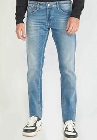 Jean bleu clair à coupe slim, avec cinq poches, une fermeture à boutons et une texture délavée, porté avec des baskets noires et blanches.