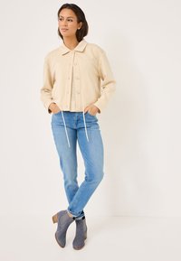 Beige kort jacka med knäppning och krage, kombinerad med ljusblå jeans och grå mocka stövlar med en träklack.