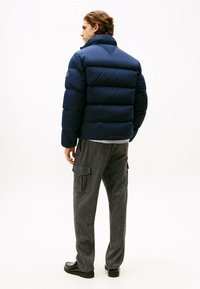 Veste matelassée navy avec un col haut, présentant un quilting horizontal et un logo sur l'épaule gauche. Associée à un pantalon cargo gris.