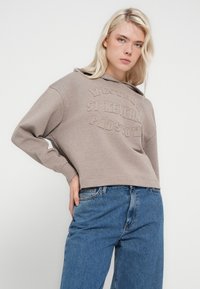 Kavbojka rjave barve z žametnim reliefnim logotipom, z kapuco z vrvico in širokimi rokavi, kombinirana s svetlo modrimi kavbojkami s pravimi hlačnicami.