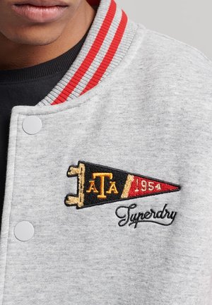 Primer plano de una chaqueta universitaria gris con cuello a rayas rojas, botones de presión blancos, un banderín bordado que dice "ATA 1954" y el logo de "Superdry".
