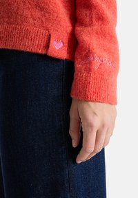 Leuchtend orangefarbener Strickpullover mit gerippten Bündchen, rosa Herz und Logo-Stickerei am Saum. Über dunklem Denim-Stoff getragen.