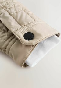 Beige quiltet jakkeærme med sort trykknap over hvid ribbet manchet på en lys baggrund.