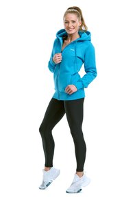 Tirkizna zip-up dukserica s kapuljačom, prednjim džepovima i vezicama, uparena s crnim tajicama i bijelim sportskim cipelama. Gladak materijal.
