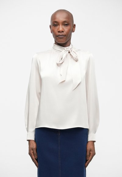 Blouse beige clair avec un nœud au cou, manches longues et texture lisse ; assortie à une jupe bleu foncé.