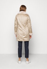 Abrigo beige de aspecto ante con un amplio cuello y puños de piel, con un corte entallado y detalles de costuras verticales, llevado sobre vaqueros oscuros y zapatillas.