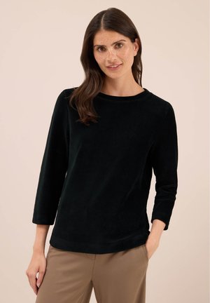 Femme aux longs cheveux bruns portant un haut noir à manches longues et un pantalon beige, se tenant avec une main dans la poche devant un fond uni.