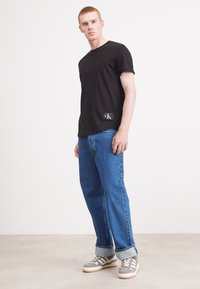 Black cotton t-shirt with short sleeves, featuring a logo patch on the lower left. Worn with loose blue denim jeans and gray shoes.