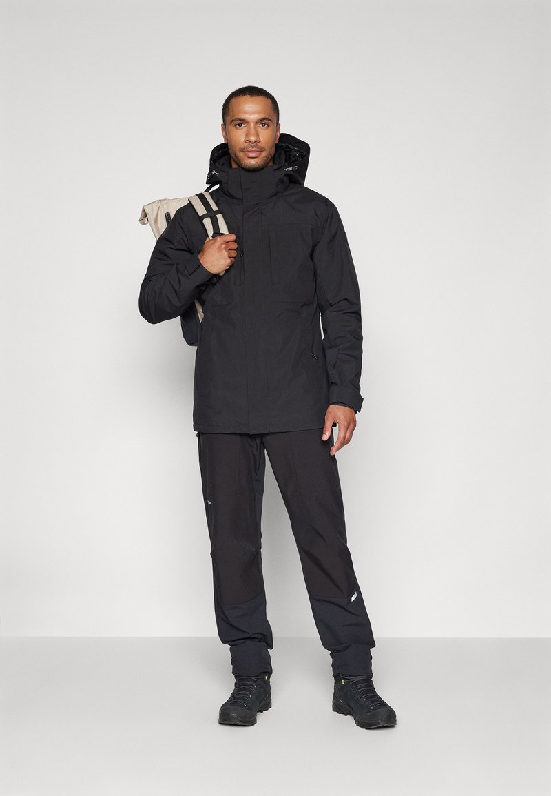 Schwarze wasserdichte Jacke mit hohem Kragen, Reißverschluss und zwei Brusttaschen, kombiniert mit passenden schwarzen Hosen und Wanderschuhen.