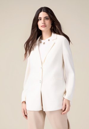 Blazer bianco con design sartoriale, rever a punta, chiusura con un solo bottone e tasche laterali, indossato sopra una maglietta chiara e pantaloni color crema.