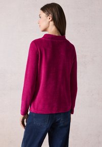 Pullover fucsia in tessuto morbido e testurizzato, con maniche lunghe e colletto. Presenta un orlo dritto e spacchi laterali. Indossato con jeans blu scuri.