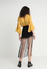 Haut jaune transparent à manches cloche associé à une mini-jupe noire avec des détails en frange, complété par des bottines à boucles.
