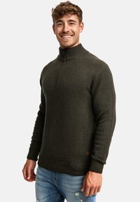 Suéter de punto verde oscuro con cuello de media cremallera, puños y dobladillo acanalados. Combinado con unos vaqueros claros desgastados.