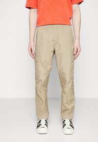 Beiga cargobyxor med en avslappnad passform, som har två sidofickor och ett texturerat tyg. Kombinerat med en klar orange t-shirt och mönstrade sneakers.