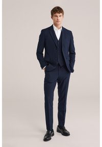 Mann steht mit den Händen in den Taschen und trägt einen marineblauen Dreiteiler, ein weißes Hemd und schwarze Anzugschuhe vor einem einfarbigen Hintergrund.