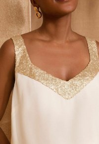 Bílý bezrukávový top s výstřihem do V ozdobený zlatými flitry. Hladká tkanina s jemným leskem. Kulaté zlaté náušnice.