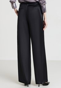 Eine Frau trägt schwarze weite Hosen, eine graue Satinbluse und schwarze Kitten-Heel-Schuhe und steht vor einem schlichten hellen Hintergrund.