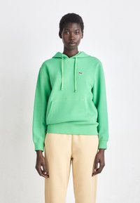 Felpa verde in misto cotone, con tasca frontale, cappuccio con coulisse e piccolo logo sul petto, abbinata a pantaloni beige.
