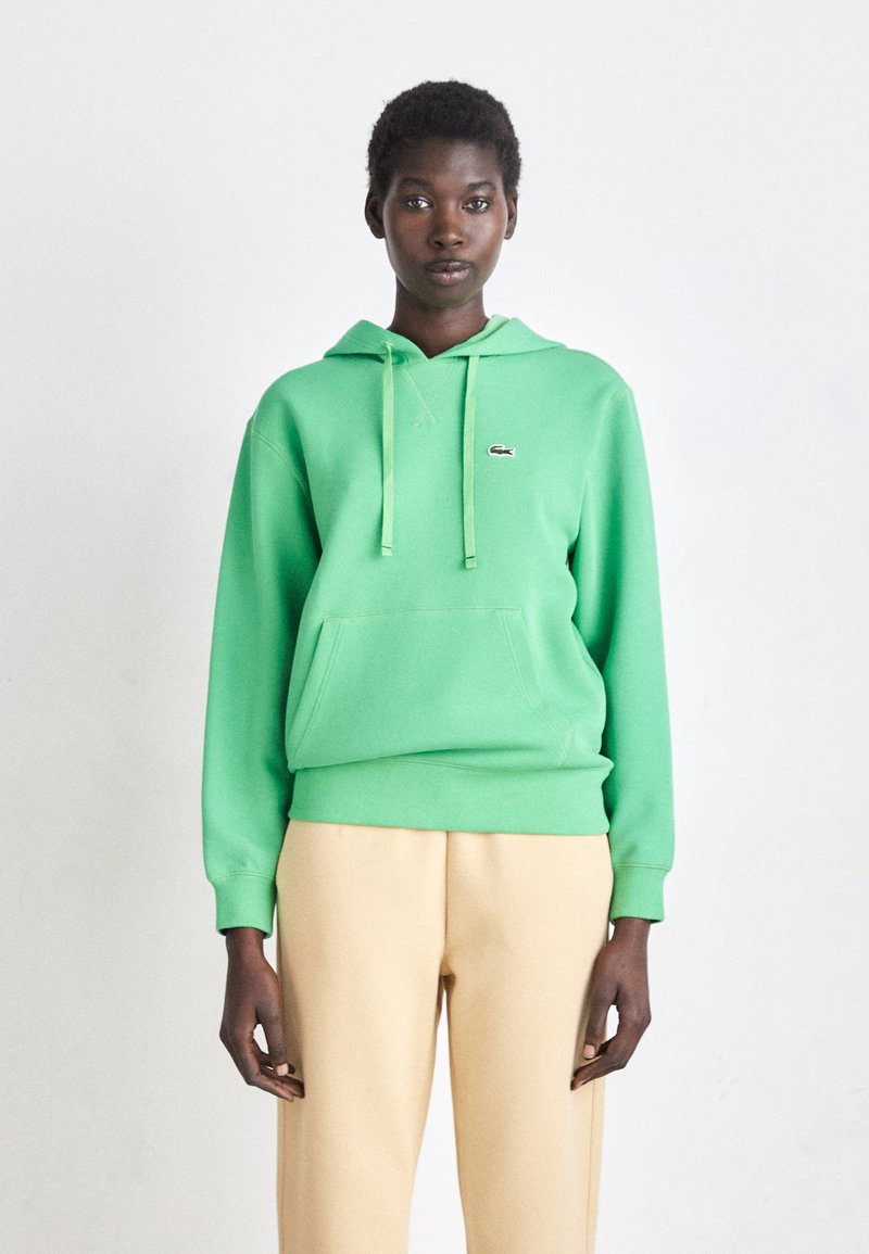 Felpa verde in misto cotone, con tasca frontale, cappuccio con coulisse e piccolo logo sul petto, abbinata a pantaloni beige.