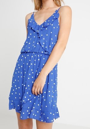 Blue sleeveless dress with white polka dots, ruffled V-neckline, elastic waist, and ruffled hem worn by a standing woman.