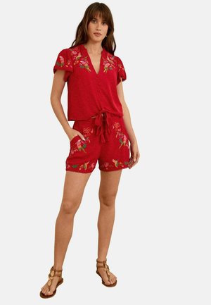 Rød to-delt sæt bestående af en kortærmet top med knapper og bindefront, parret med broderede shorts og brune sandaler.