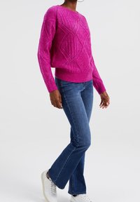 Fuchsiový pletený svetr s diamantovými vzory, širokými manžetami a volným střihem, v kombinaci s modrými zvonovými džíny a bílými teniskami.
