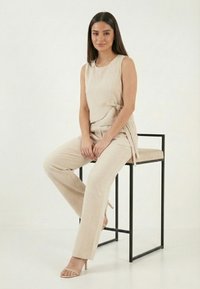 Mujer con cabello largo y oscuro sentada en un taburete de metal negro minimalista, vestida con una blusa sin mangas beige y pantalones a juego, con sandalias de tacón alto.