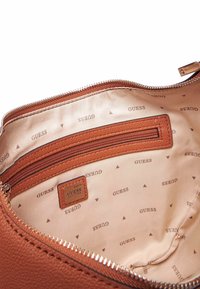 Interno di una borsa a mano in pelle marrone testurizzata con fodera in tessuto che presenta il logo ripetuto "GUESS" e piccoli triangoli. Include una tasca con zip.