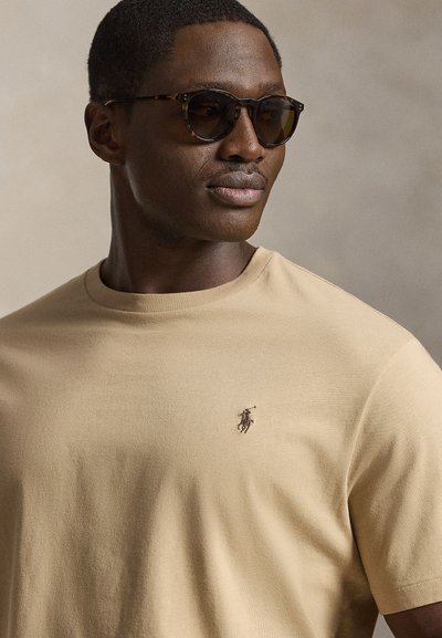 Mann mit Hornbrillengestell und beigem T-Shirt mit kleinem gesticktem Polo-Spieler-Logo auf der Brust.