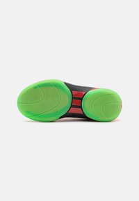 Semelle de chaussure en noir, en matériau texturé avec un motif en caoutchouc vert et rouge, présentant des rainures circulaires et deux sections circulaires vertes.