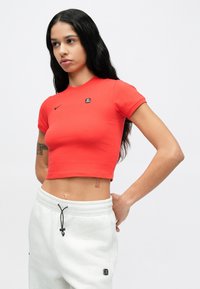 Červený crop top vyrobený z hladkej látky, s malým čiernym logom Nike a krátkymi rukávmi, doplnený svetlosivými teplákmi.