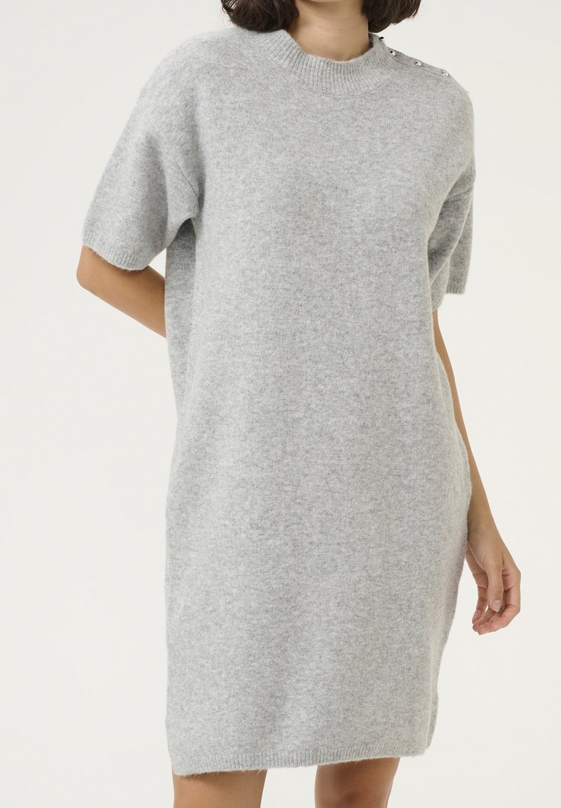 Femme portant une robe-pull en maille gris doux à manches courtes avec des détails de boutons sur l'épaule, debout devant un fond uni.
