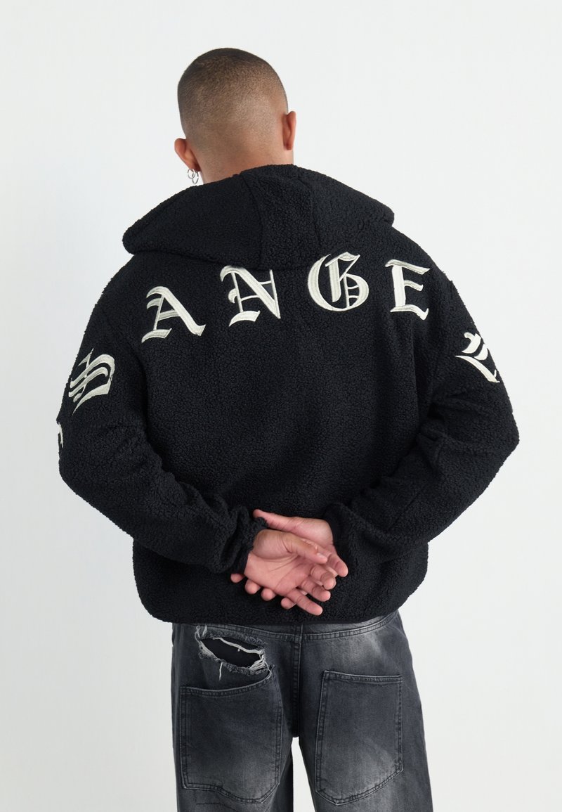 Homme aux cheveux courts et boucles d'oreilles portant un pull à capuche noir avec des lettres gothiques blanches dans le dos et un jean gris délavé avec une déchirure.