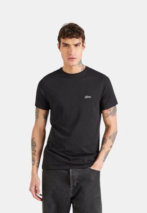 Uomo con tatuaggi che indossa una t-shirt Guess nera a maniche corte e jeans scuri, in piedi con una mano in tasca contro uno sfondo bianco.