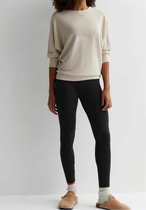 Femme portant un pull beige à rayures, des leggings noirs, des chaussettes blanches et des chaussures à enfiler couleur beige, debout devant un fond clair uni.
