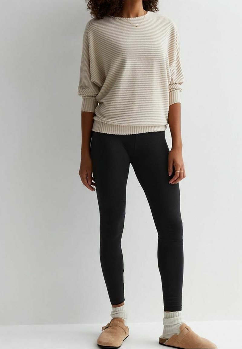 Femme portant un pull beige à rayures, des leggings noirs, des chaussettes blanches et des chaussures à enfiler couleur beige, debout devant un fond clair uni.