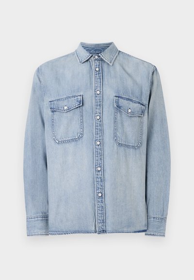 Cămașă din denim bleu deschis, cu mâneci lungi, guler ascuțit, închidere cu nasturi în față, două buzunare pe piept cu clapete și detalii vizibile de cusătură.