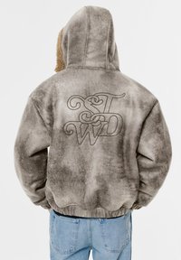 Sweat à capuche en polaire grise avec une finition texturée et déchirée. Présente un grand logo brodé au dos et une doublure de capuche contrastante en peluche.