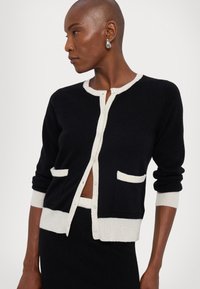 Black cardigan with cream trim, featuring a round neckline, button closure, and two front pockets. Soft knit texture, close-fitting design.