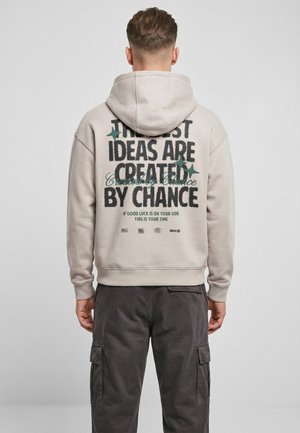 Mann trägt einen hellbeigen Hoodie mit dem Text "The best ideas are created by chance" und dunkle Cargohosen, steht vor einem schlichten Hintergrund.