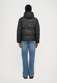 Schwarze Daunenjacke mit Kapuze und auffälligem, einheitlichem Branding. Der Stoff hat eine glatte Textur und ein gestepptes Design. Kombiniert mit blauen Jeans.