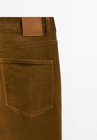 Pantalon en velours côtelé marron avec une étiquette en cuir sur la ceinture et une poche arrière présentant des détails de couture visibles et des nervures verticales.